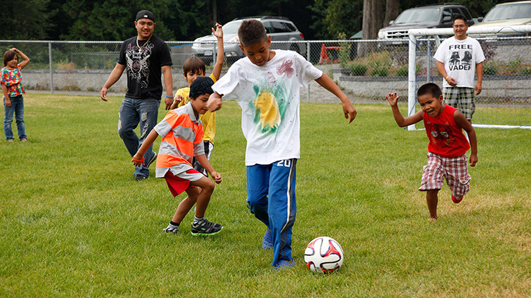 Young boys and several men are playing soccer on a grass field. One of the younger boys has possession of the ball near the center of the image.