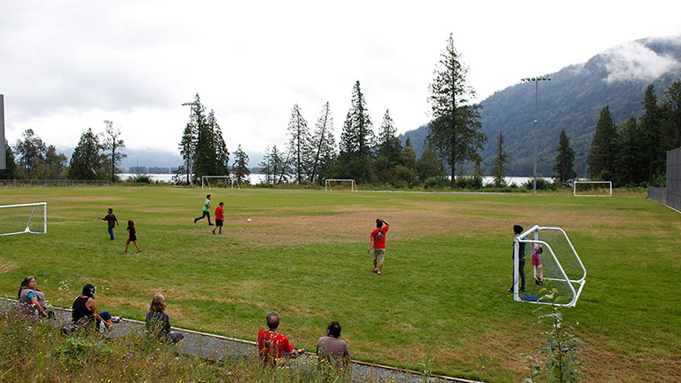 Une partie de soccer sur un terrain extérieur gazonné. À l’arrière-plan, il y a des arbres et des montagnes. Au premier plan, plusieurs spectateurs regardent la partie, appuyés sur une clôture.