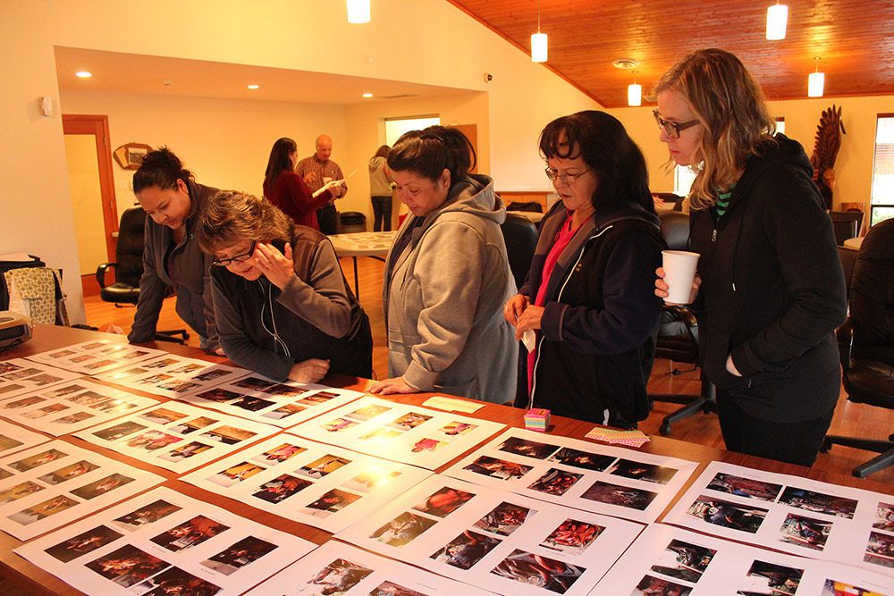 Five people are gathered around a long rectangular table, leaning forwards and looking at many pages of photographs laid out on the table in front of them.