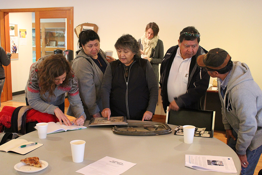 Five people are gathered around a circular table, looking at different objects including pictures and books and engaging in conversation.