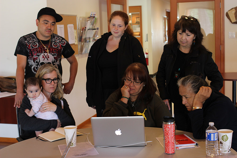 Six people are gathered around a table, looking at a laptop computer screen. Three people are standing, and three people are sitting in front of them. The woman sitting on the left side is holding a baby in her arms.