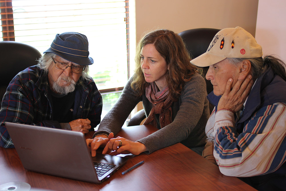 Three people are gathered at a table, sitting, and looking earnestly at a laptop computer screen. The woman sitting in the middle is typing, while the men sitting on either side are looking at the screen.