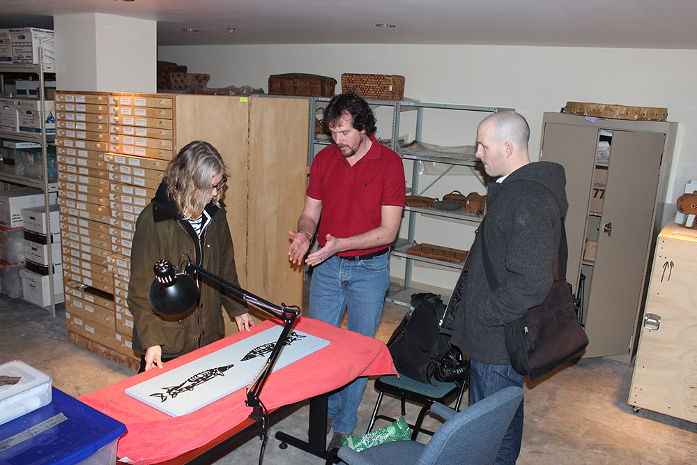 Trois personnes debout autour d’une petite table regardent une peinture posée sur un tissu orange. Sur la peinture, deux esturgeons noirs se font face sur un fond blanc.