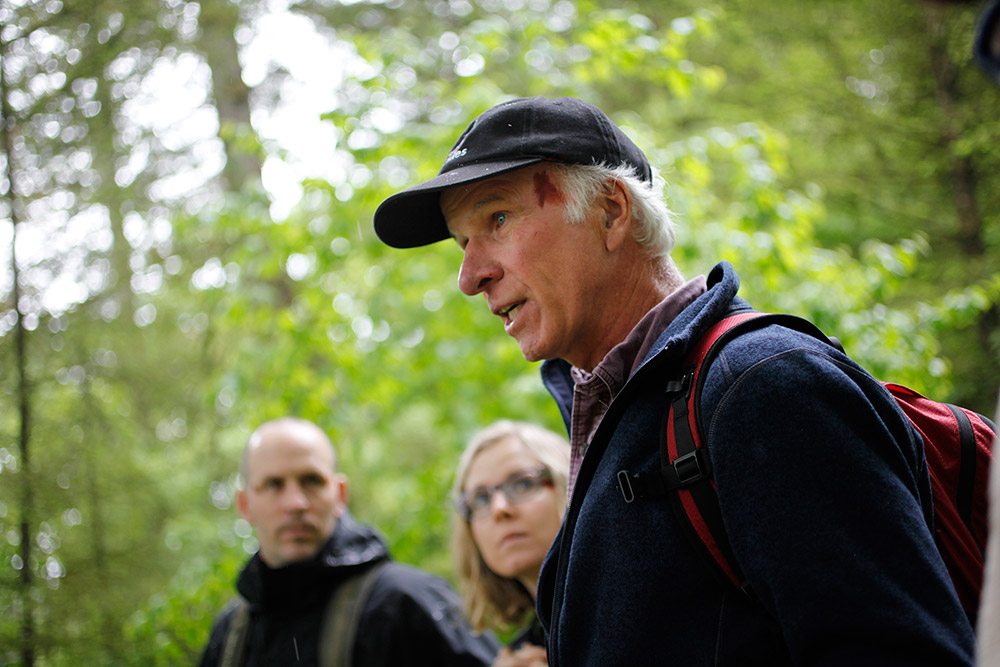 Three people are walking through a forested area; the man in the foreground is talking, while the other man and woman are listening.