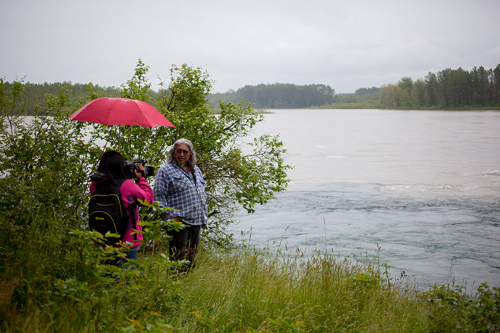 A man stands by the edge of the water, looking back over his shoulder towards the trees as he is filmed by a woman holding a red umbrella.