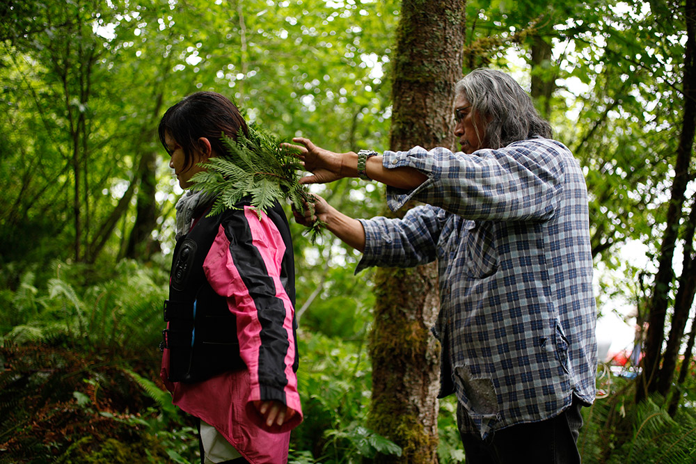 A man stands on the right side of the photo, with his arms outstretched. He is holding a cedar bough over the shoulders of a woman, who is standing with her back to him. 