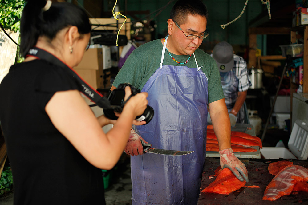 A woman is reviewing a photo on her camera in the foreground, and a man stands in front of her looking down at the salmon he is filleting. He is wearing a blue apron, and holds a knife in one hand.