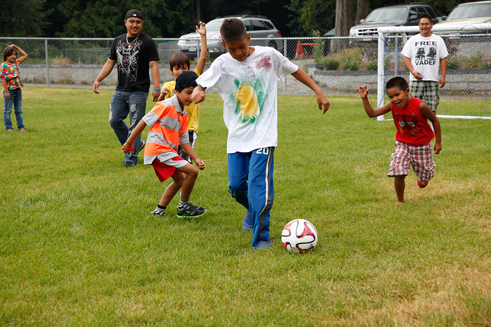 Young boys and several men are playing soccer on a grass field. One of the younger boys has possession of the ball near the center of the image.