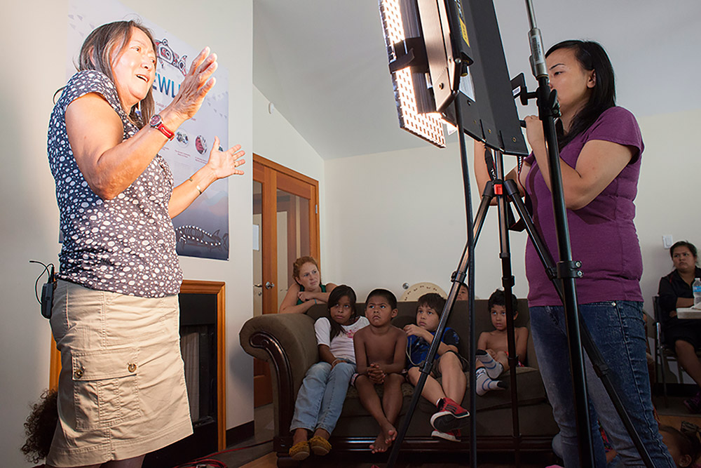 A woman is standing on the left side of the photo, sharing a story with children that are seated on a couch in the background. There is a woman on the right side that is filming the event.