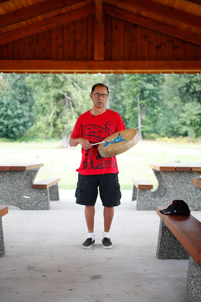 A man stands outdoors playing a hand drum. The drum is decorated with the image of a blue hummingbird.