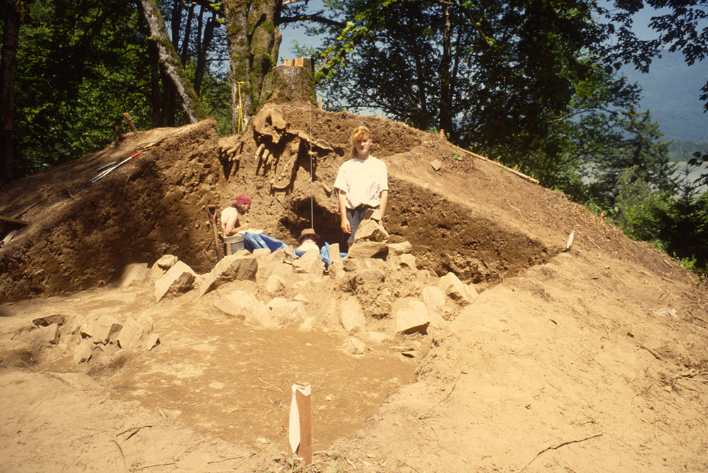 Several people are gathered alongside an excavated large mound of earth. The mound is 3 m tall and has been sectioned in half lengthwise. There are many large rocks towards the base of the mound.