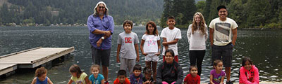 A group of children and young adults pose with their elder for a photograph on the shore of the river. There are mountains and trees in the background.