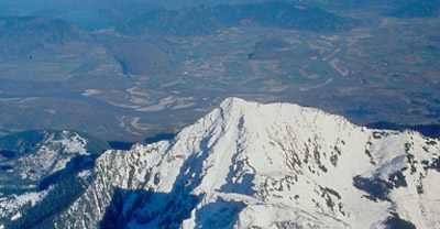 Aerial view of a snow-capped mountain with a green mountainous landscape in the background.