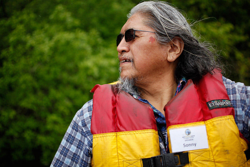 A man drives a small motorboat across the river. He wears a red and yellow lifejacket.