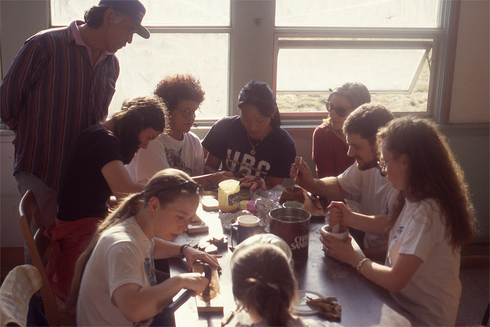 Many students are gathered at a rectangular table, using various tools and materials, including red ochre.