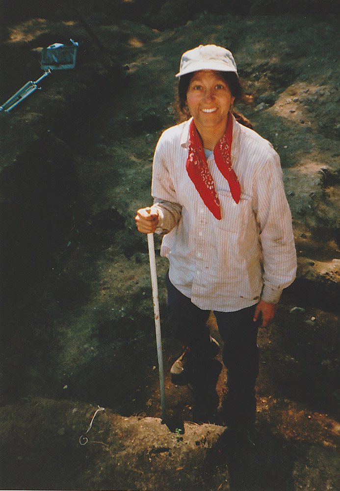 A woman with a red kerchief stands in a section of dirt as she excavates the area around her.