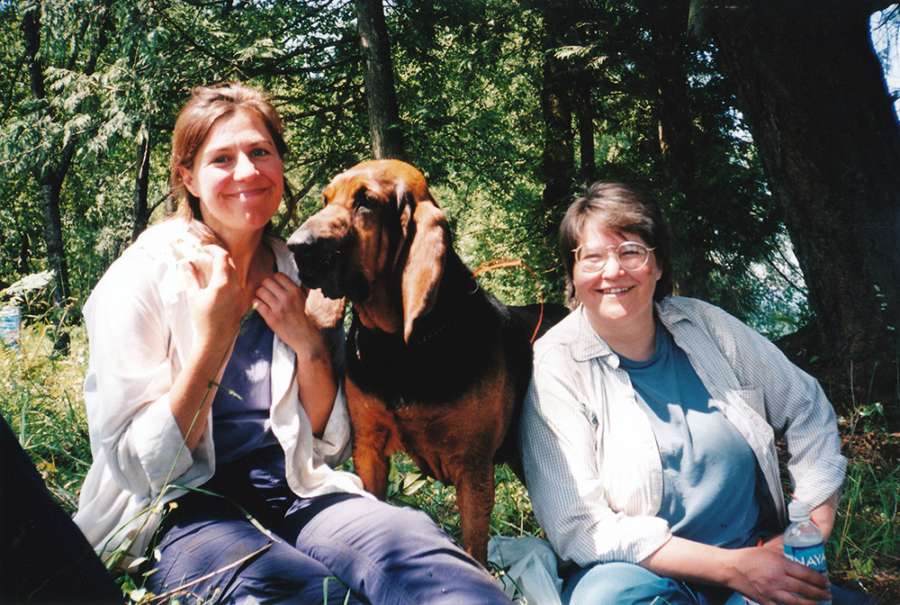 Two women sit on the grass, with a dog standing between them.