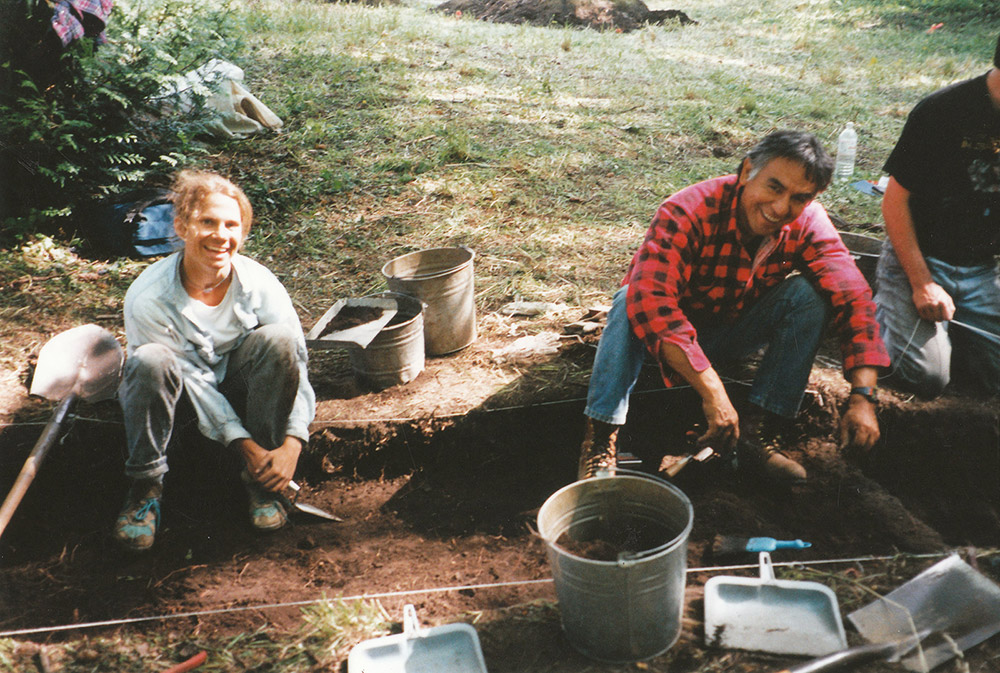 Two people sit within a sectioned archaeological area, using shovels to excavate the earth.