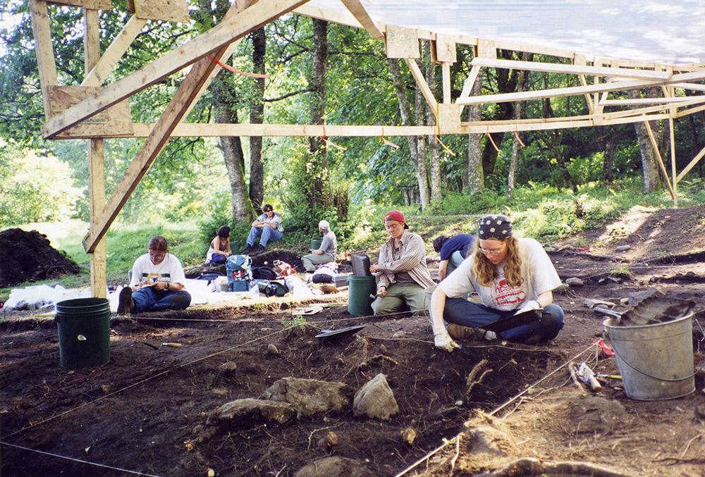 A group of archaeologists excavate the earth in sections; they are underneath a rain shelter.