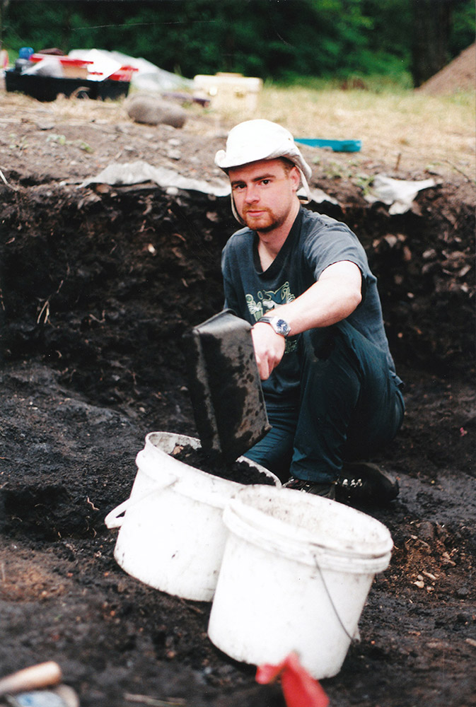 A young man kneels within an excavated section of earth, shovelling dirt into a bucket.