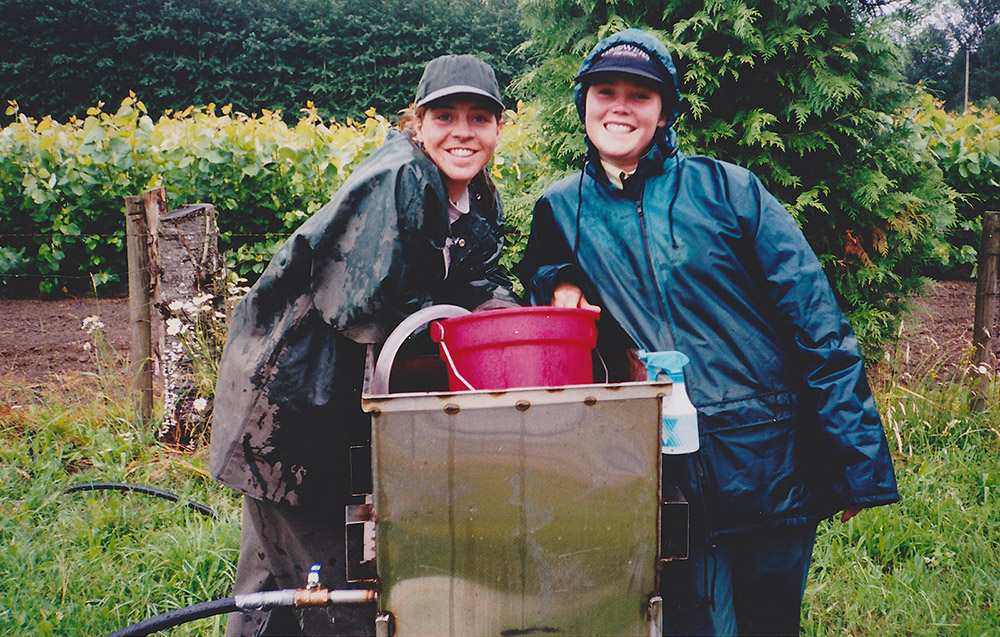 Two women stand behind a large container of plant remains, using a bucket to sift through its contents.