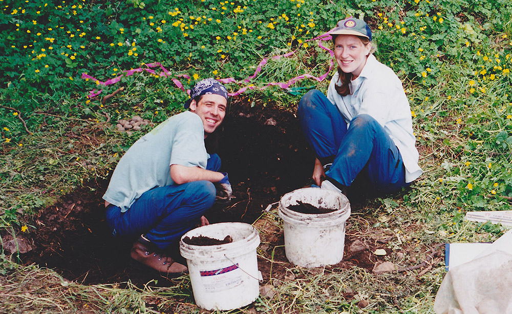 A man and woman sit in a small excavation site, with buckets of dirt by their feet.
