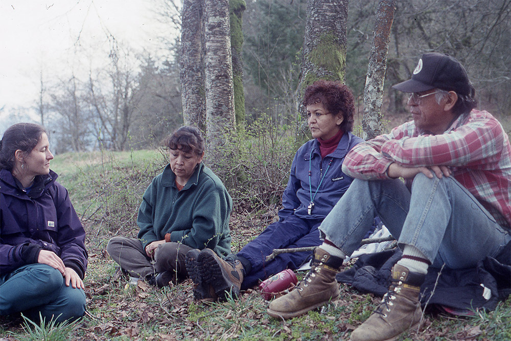 A woman listens to the stories of three elders. All are seated together on the grass.