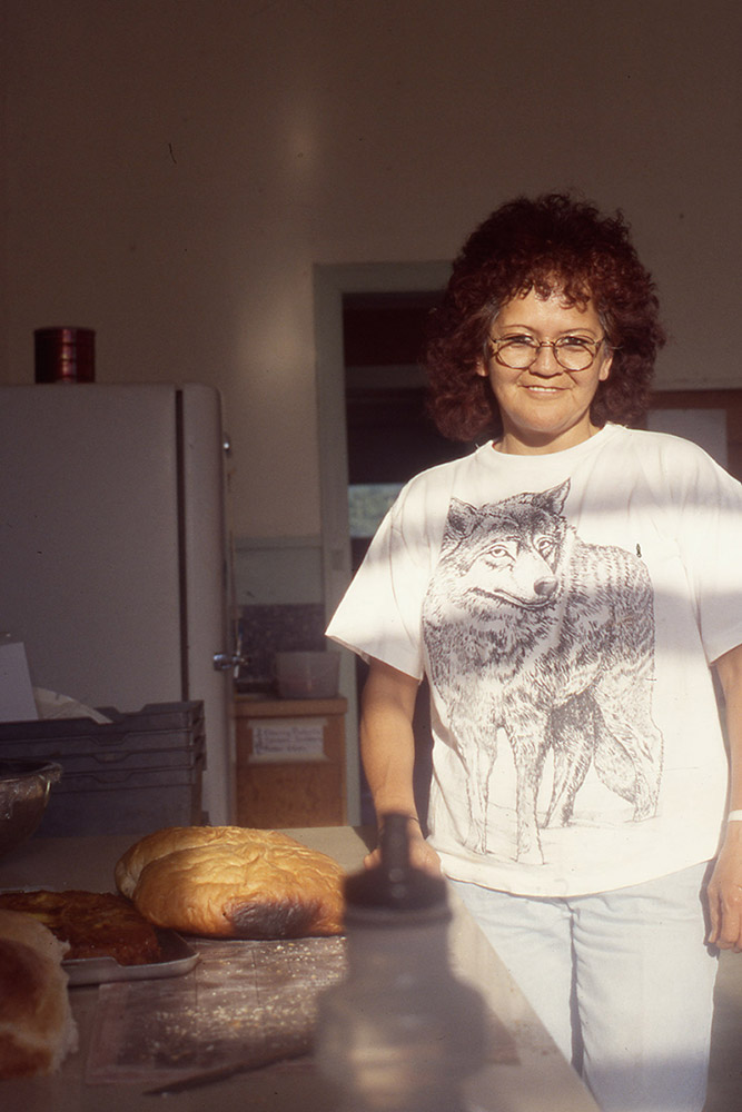 A woman stands beside a kitchen counter; she is smiling. There is freshly baked bread on the table.