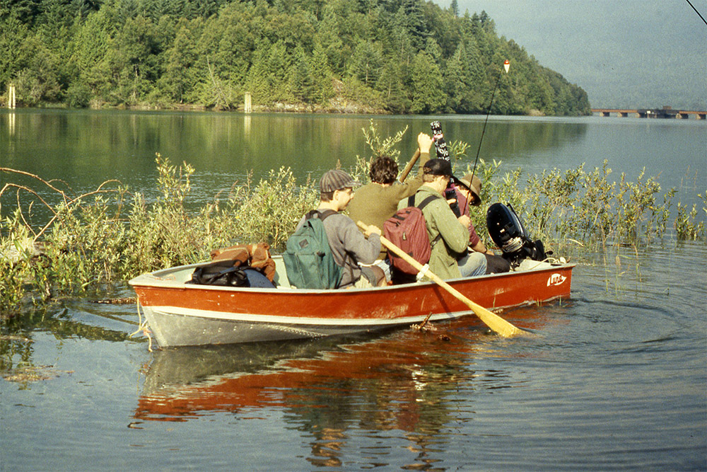 Quatre hommes assis dans un petit bateau à moteur sur la rivière. L’homme assis près de la proue tient une rame.