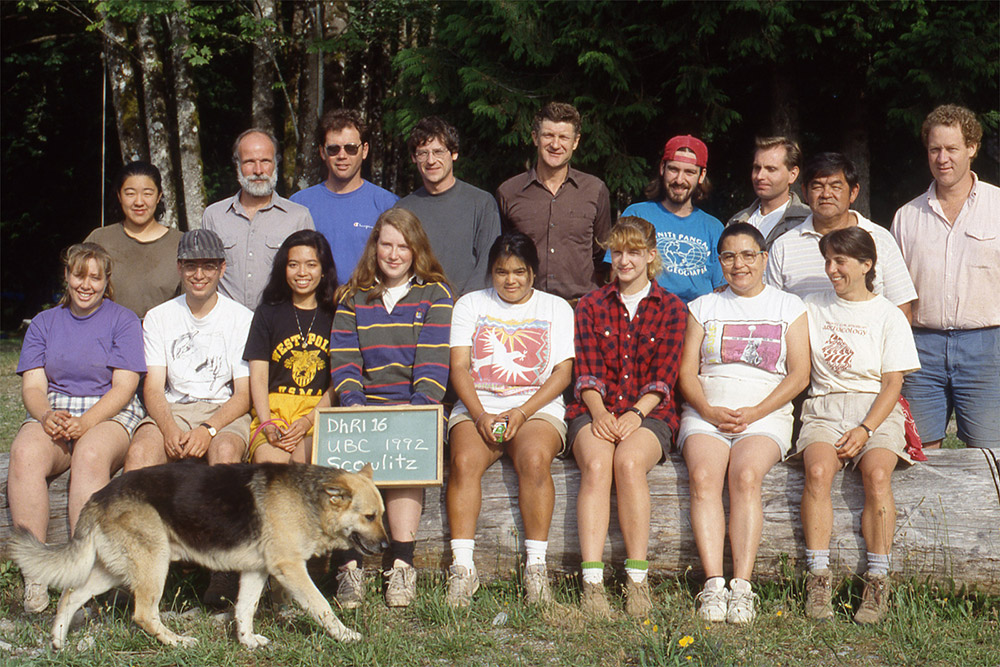 Two rows of people pose for a photograph. The front row is seated, and the back row is standing. The front-middle student holds a board with the class/year information. A dog walks in the foreground of the picture. 