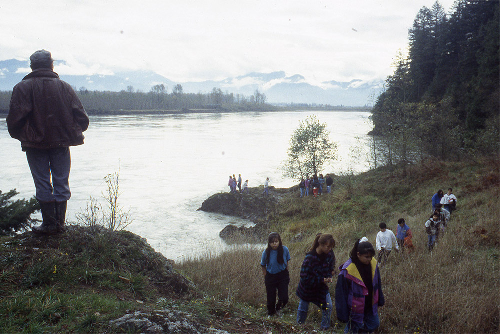 A group of people hike along the grassy shoreline; a man stands at a higher elevation looking back towards the river and mountains.