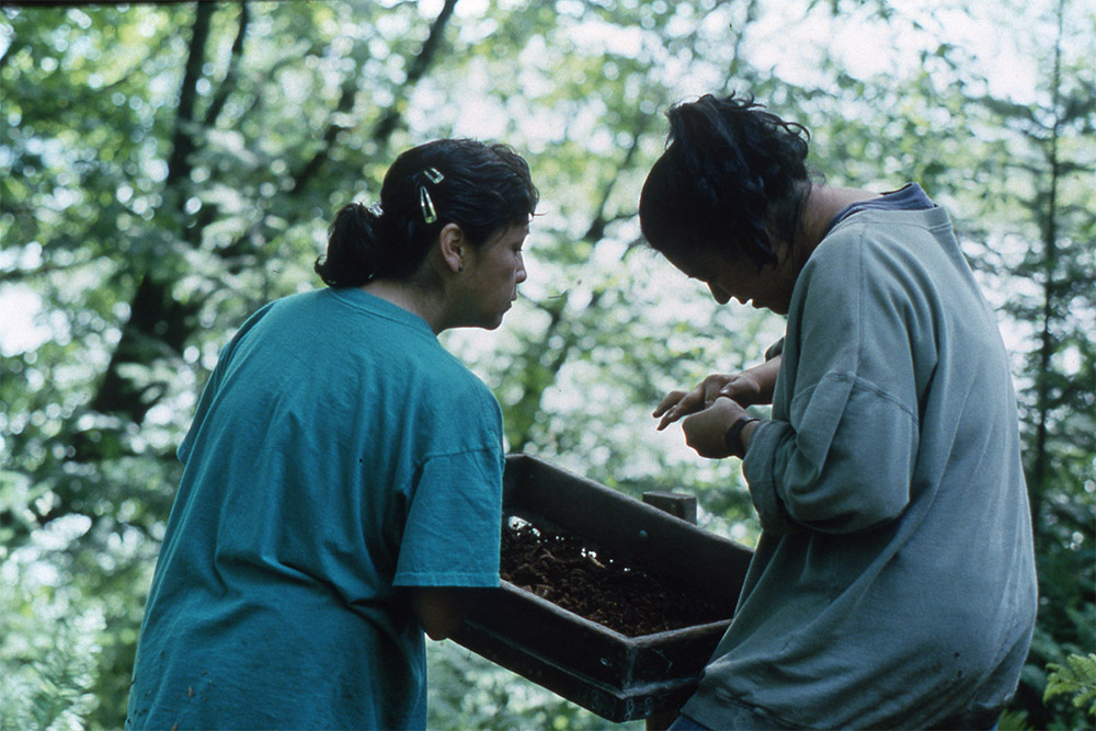 Two women hold up a wooden screen, sifting through dirt and examining the contents up close.