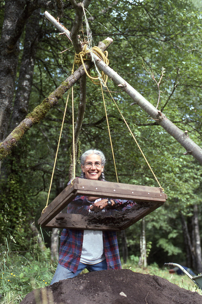 A woman leans over a wood-framed screen, as she sifts through the contents. The screen is tied with rope to the wooden structure above, and a large mound of dirt sits underneath.
