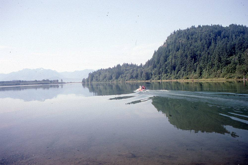 A group of people travel across the Harrison River by motorboat. The water is calm, and there are trees and mountains in the background.