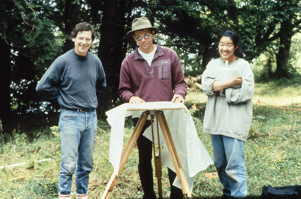 Three people stand outside around a small table, with a piece of fabric draped over it.