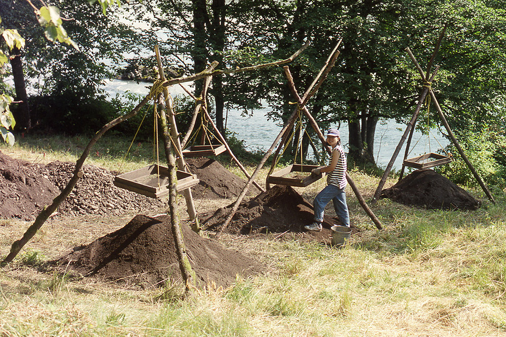 A woman stands beside a hanging wooden screen, sifting through dirt into a large mound below. There are many other hanging screens and dirt mounds set up around her.