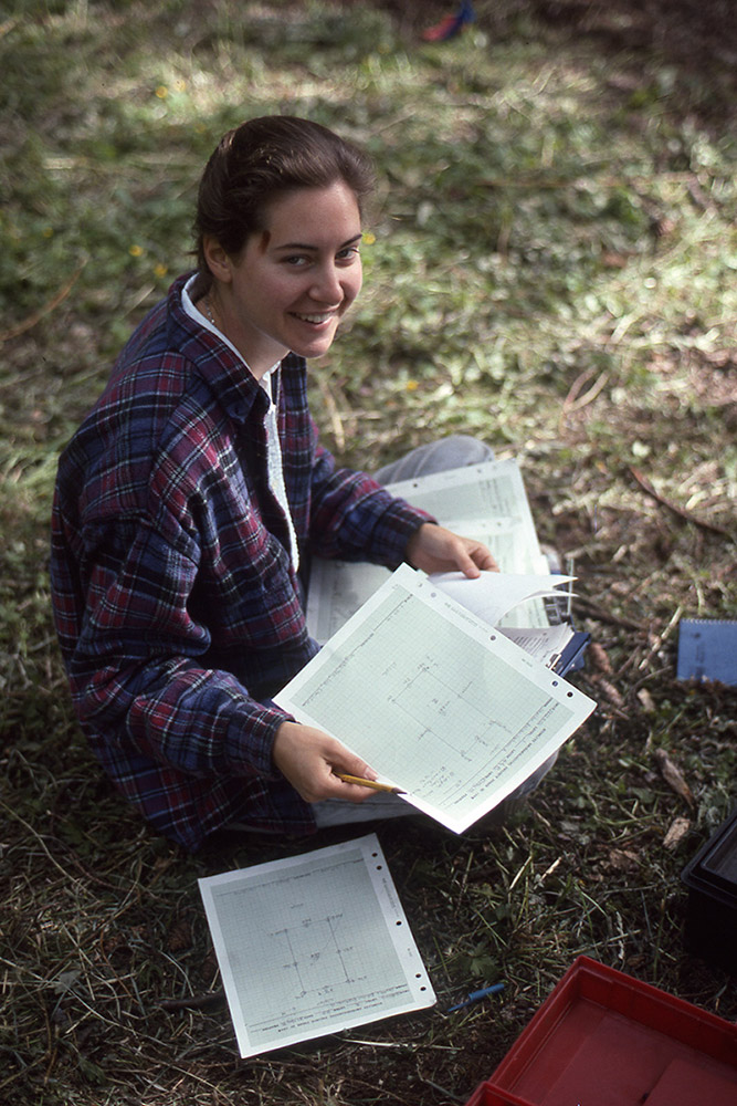 A woman sits on the grass and sorts through a pile of papers on her lap. She is smiling.