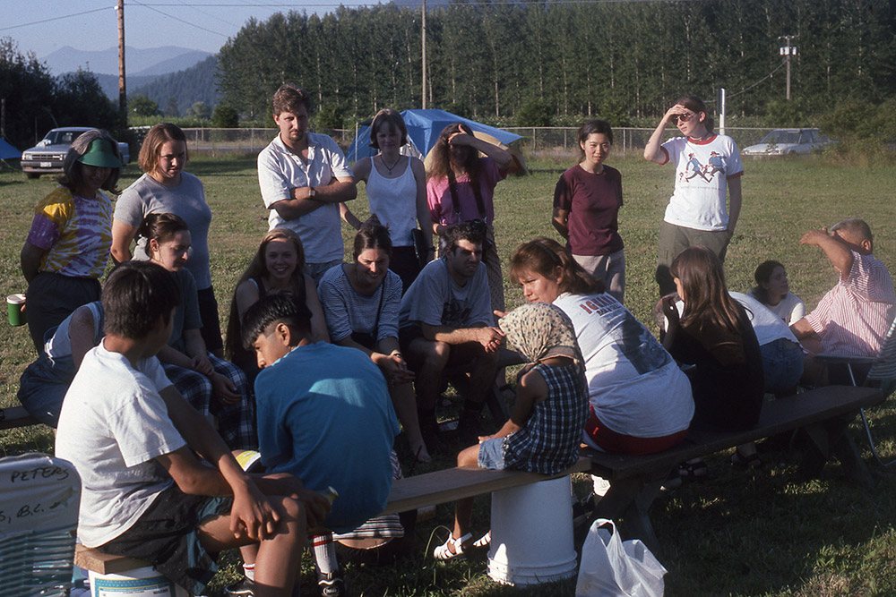 A group of students are gathered outside on a bench on a grass field, while a woman in the center explains the rules of a game.