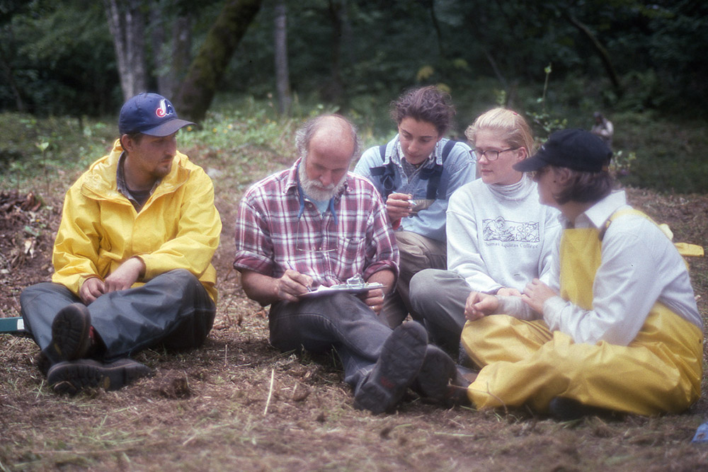 Four students are seated on the grass beside their teacher, listening to his lecture.