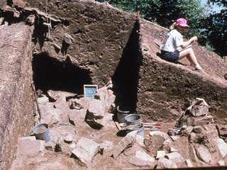 Une femme assise à côté d’une section de terre qui a été creusée. À l’intérieur de la section se trouve un gros tas de pierres.