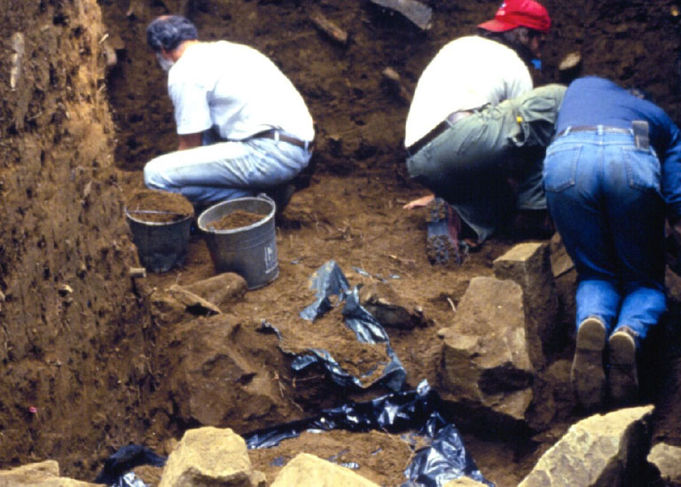 Plusieurs personnes travaillent à l’intérieur d’un tertre ancien qui a été creusé. Elles sont entourées de murs de terre, de roches et de seaux remplis de terre.