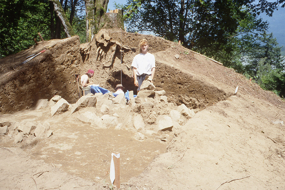 Several people are gathered alongside an excavated large mound of earth. The mound is 3 m tall and has been sectioned in half lengthwise. There are many large rocks towards the base of the mound.
