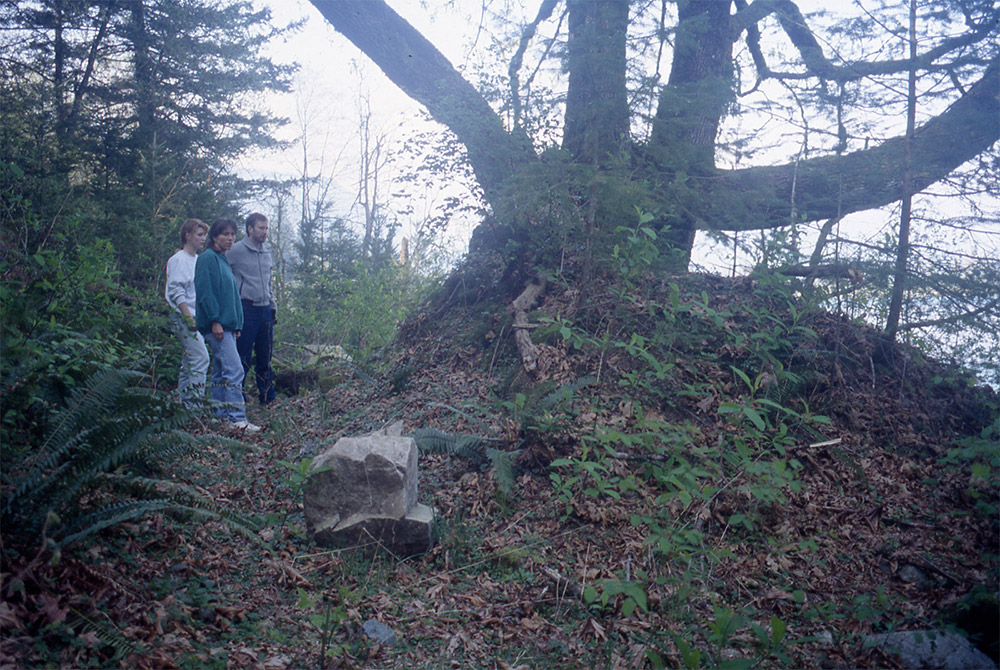 Three people stand beside an earthen mound with a large maple growing out of it.