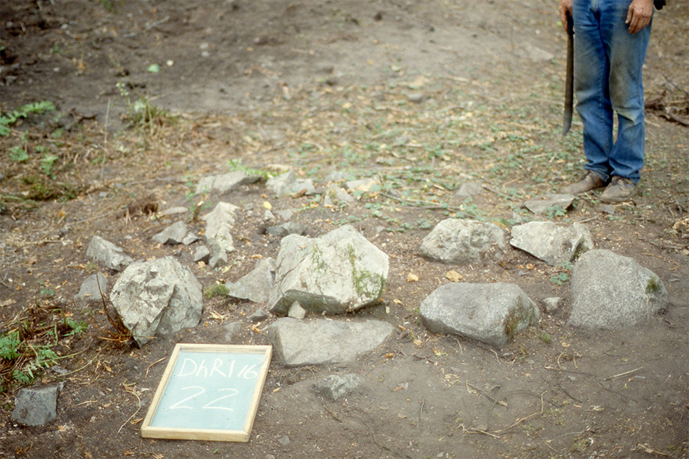 A small chalkboard is placed beside a pile of large rocks; a person stands in the background holding a machete.