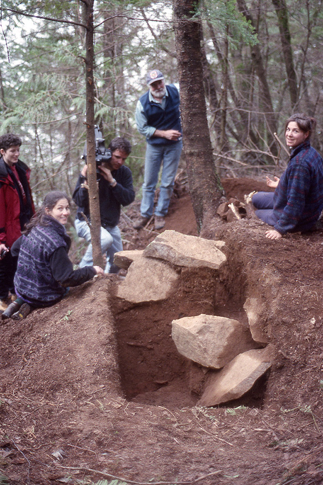 A group of archaeologists sit beside a section of earth that contains large boulders.