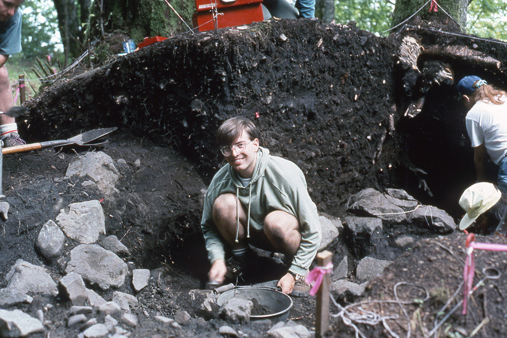 A young man man sits in an archaeological area and is shoveling dirt into a bucket.