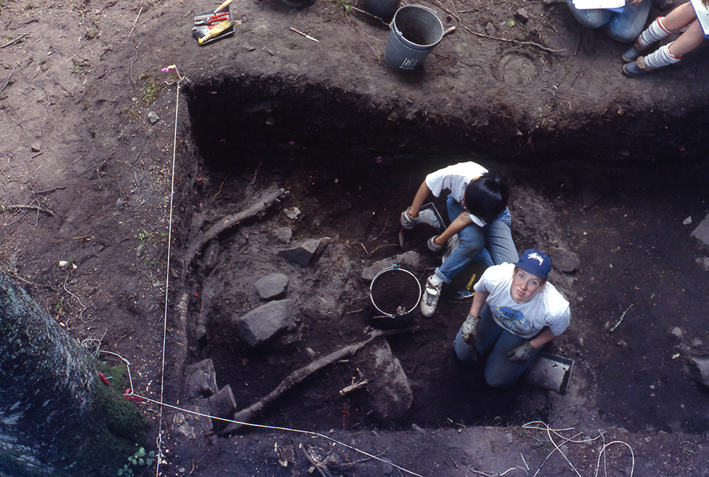 Two archaeologists sit in the section of earth they are excavating. The woman on the right is looking up at the camera.