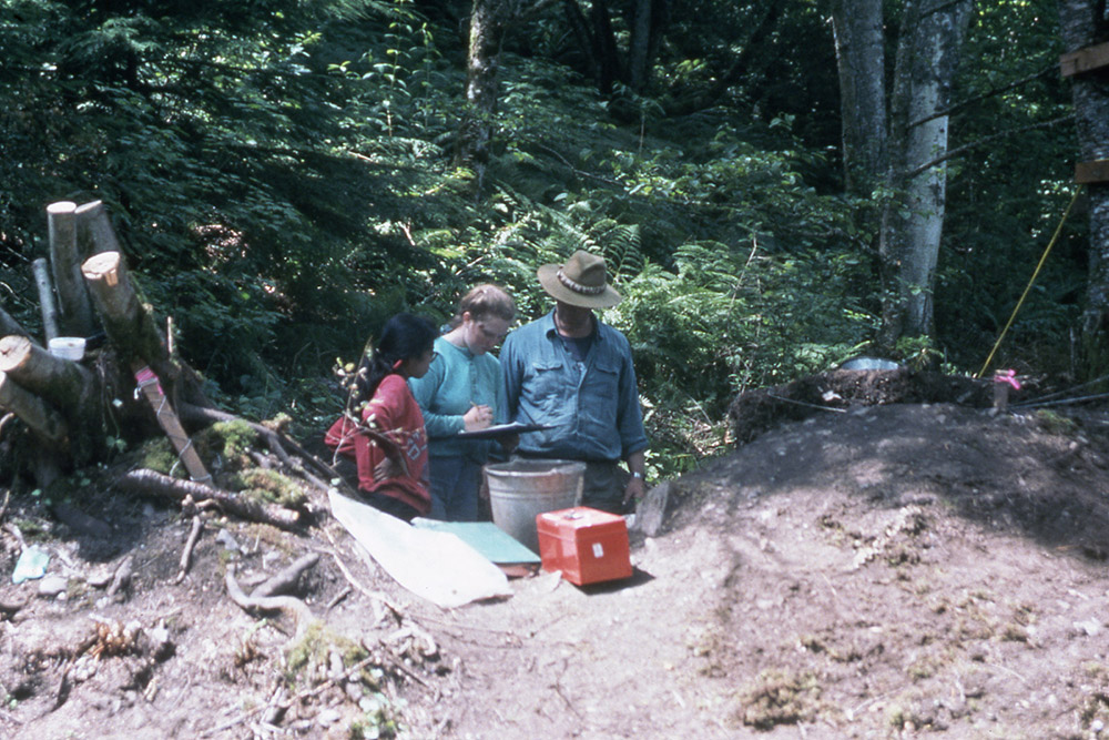 Three archaeologists stand beside each other in conversation; the woman in the middle is taking notes.
