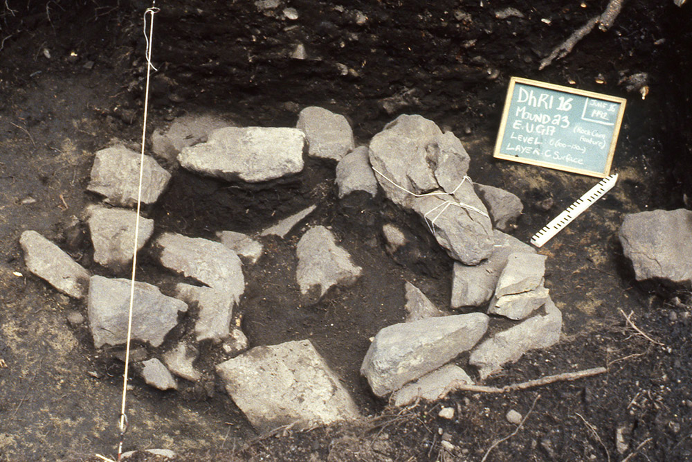 A chalkboard and ruler are placed beside a circular pile of large rocks inside an excavated mound.