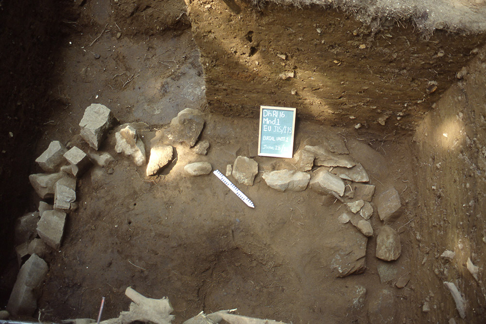 Inside an excavated mound, a chalkboard and ruler are placed beside a square shaped pile of large rocks.